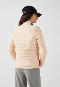 Giacca imbottita rosa chiaro con design a righe orizzontali. Presenta un collo rotondo e una silhouette aderente. Indossata con un cappellino nero.