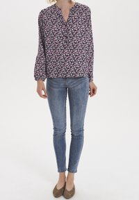 Blouse fleurie à manches longues, col en V, fond noir, fleurs roses et blanches. Associée à un jean skinny bleu et des ballerines marron.
