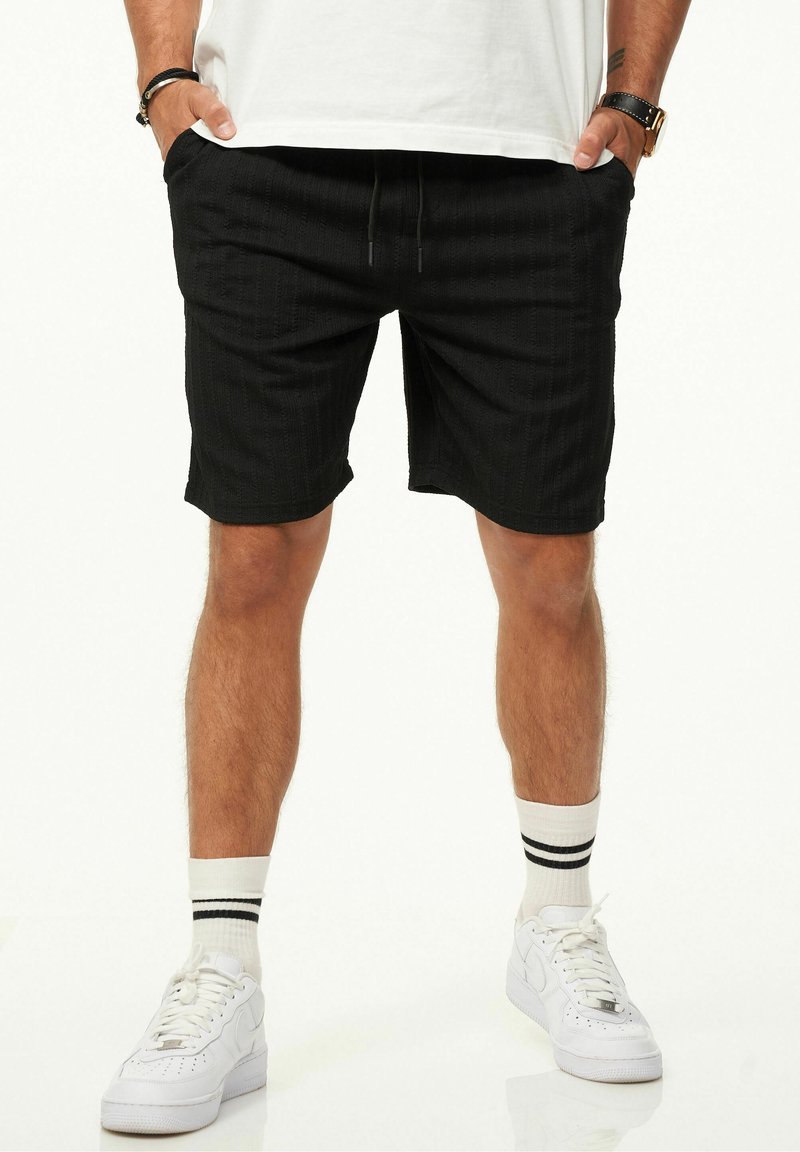 Black textured shorts with an elastic waistband and drawstring, featuring side pockets. Paired with white sneakers and striped socks.