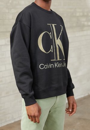 Person trägt schwarzen Calvin Klein Sweatshirt mit großem goldenem "CK"-Logo und hellgrüner Hose, steht vor einer hellen Backsteinmauer.