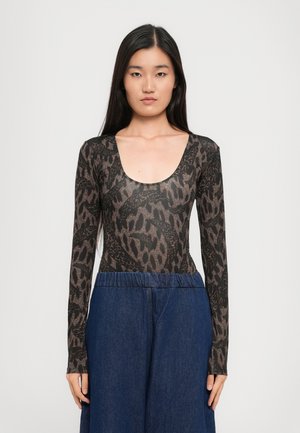Mujer con cabello largo y oscuro vistiendo una blusa ajustada de manga larga con estampado de animal y unos vaqueros azules de tiro alto y holgados, de pie contra un fondo sencillo.