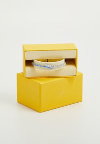 Bracelet en argent présentant une série de pierres précieuses claires et bleues, exposé sur un coussin crème à l'intérieur d'une boîte jaune.