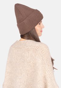 Braune Strickmütze mit geripptem Muster und spitzem oberen Ende, getragen mit einem hellbeigen gestrickten Pullover, der ein lässiges Schnittdesign präsentiert.