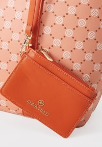 Porte-cartes zippé en cuir orange avec des ferrures dorées et un logo attaché à un fond de sac corail à motifs.