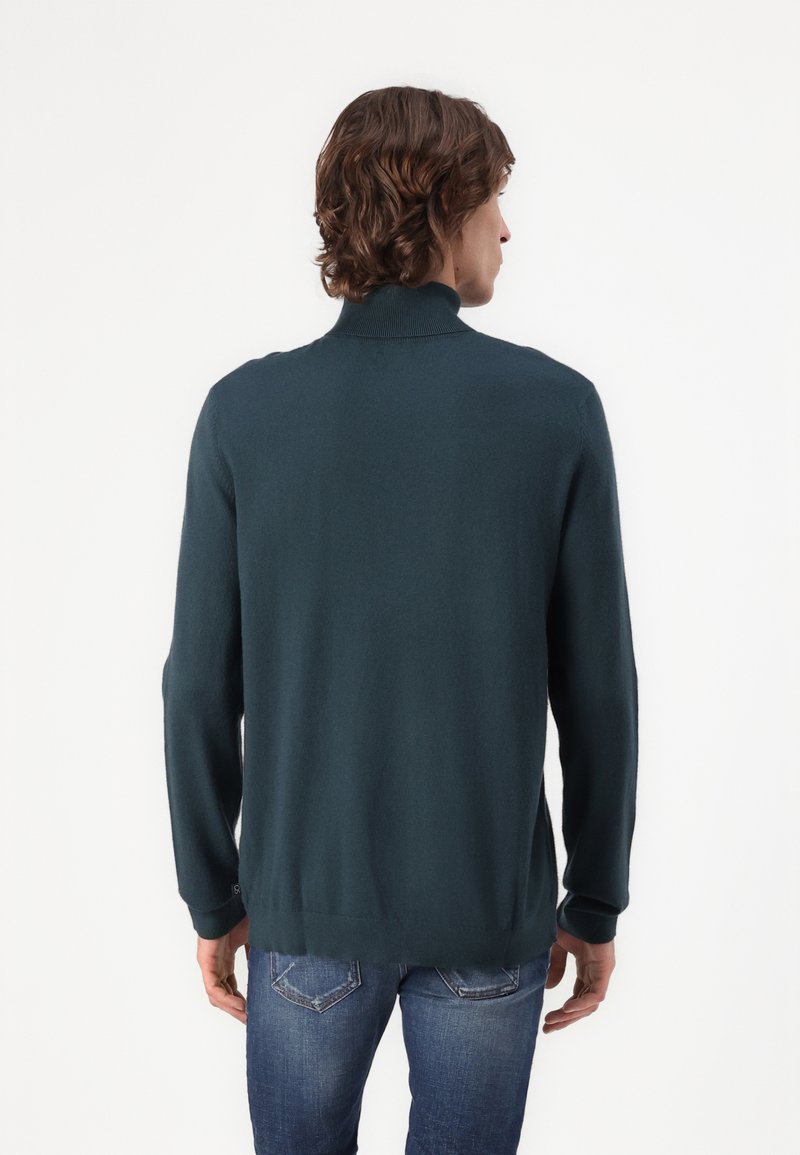 Pull en maille vert foncé avec un col roulé ajusté, des poignets côtelés et des fentes discrètes sur les côtés, porté avec un jean bleu clair. Texture lisse.