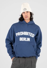 Sudadera azul marino con bolsillo frontal y texto blanco "PROHIBITED BERLIN". Gorro de punto crema con etiqueta visible. Tejido suave y casual.