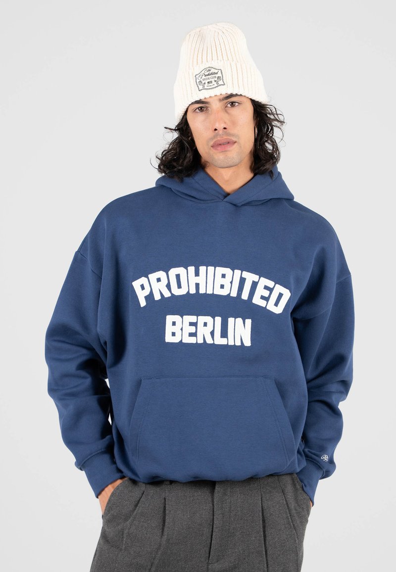 Sudadera azul marino con bolsillo frontal y texto blanco "PROHIBITED BERLIN". Gorro de punto crema con etiqueta visible. Tejido suave y casual.