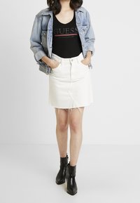 Giacca di jeans, top nero a V con logo "GUESS", gonna di jeans bianca con orlo sfrangiato e stivali neri alla caviglia con punta affilata.