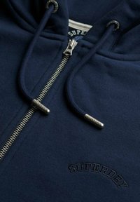 Sudadera con cremallera en azul marino, fabricada en algodón, con cremallera plateada, puntas de cordón con marca y logo bordado de "SUPERDRY" en el pecho.