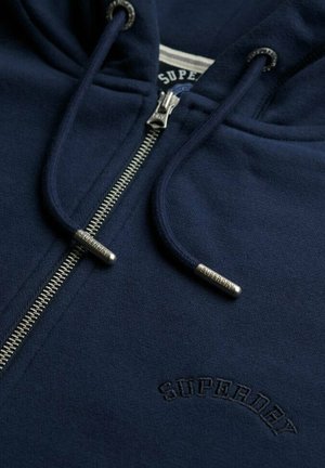Marineblaue Kapuzenjacke mit Reißverschluss aus Baumwolle, mit silbernem Reißverschluss, gebrandeten Kordelzugspitzen und gesticktem "SUPERDRY"-Logo auf der Brust.