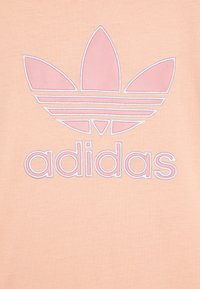 Persikų spalvos marškinėliai su rožiniu siuvinėtus Adidas logotipu, kuriame yra trefoil dizainas ir kintančios juostelės po logotipu. Medžiaga atrodo minkšta ir lygi.