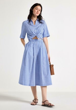 Nainen pukeutunut sinivalkoiseen ruutukuosiseen kahden osan asuun, jossa on solmittu lyhythihainen paita, midihame, ruskeat sandaalit ja hän pitää kädessään pientä ruskeaa käsilaukkua.