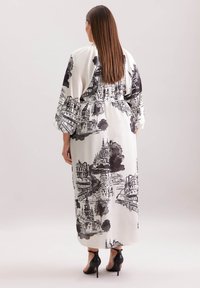 Langes weißes Kleid mit schwarzem Stadtbilddruck, breiten Ärmeln und tailliertem Bund. Schwarze High-Heels vervollständigen das Outfit.