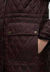 Poche matelassée marron foncé d'une veste Barbour avec boutons-pression et une main partiellement insérée sur le côté droit.