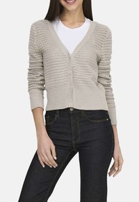 Cardigan en tricot beige clair avec un col en V, six boutons et des poignets côtelés. Présente un design perforé et associé à un jean foncé.