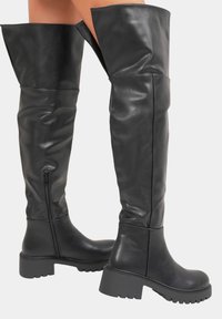 Bottes en cuir noires montantes avec des talons épais et des zips latéraux portées sur des jambes nues devant un fond uni.