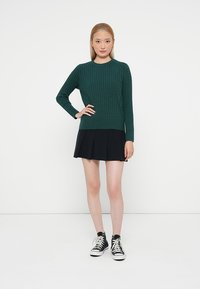 Pull en maille torsadée verte avec un col rond, associé à une jupe plissée noire et des baskets noires. Design simple et texturé.