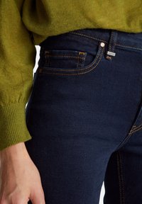 Jeans de color azul oscuro con una textura suave, costuras naranjas y un pequeño acento plateado en la cinturilla, combinados con un suéter de punto verde.