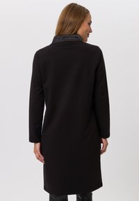 Robe noire à manches longues avec un col montant, en tissu lisse et texturé ; présente une coupe droite et une longueur sous le genou.