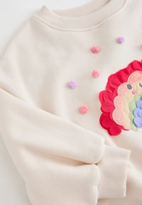 Krémově zbarvená mikina s barevným vyšívaným motivem lva a malými kulatými bambulkami v růžové a fialové barvě na přední straně.
