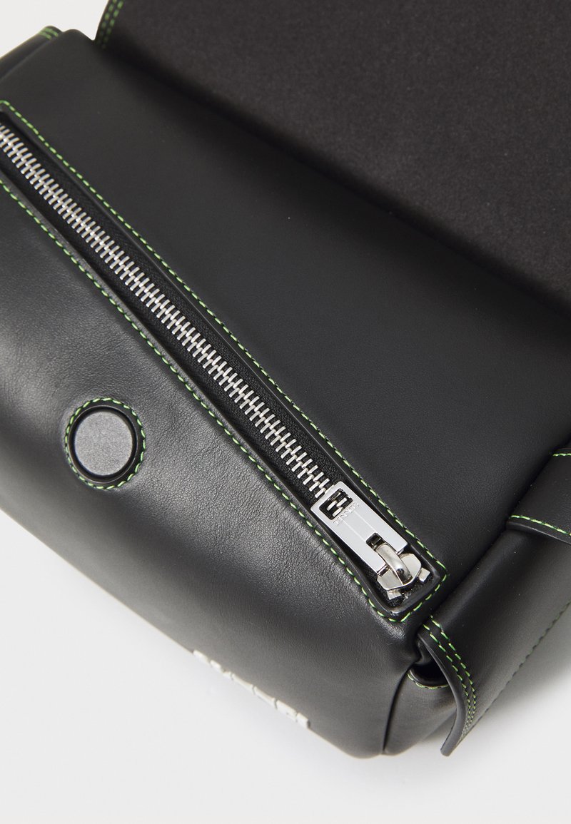 Sac en cuir noir avec une texture lisse, doté d'une fermeture éclair argentée et d'accents de couture verte. Comprend un détail en bouton circulaire.