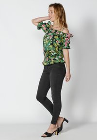 Top floreale off-the-shoulder verde, con maniche a volant e orlo peplum, abbinato a jeans skinny neri e sandali con tacco aperti davanti.