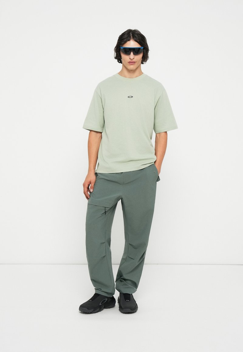 T-shirt vert clair à manches courtes avec un petit logo, associé à un pantalon vert foncé et des chaussures noires à enfiler. Texture mate sur l'ensemble.