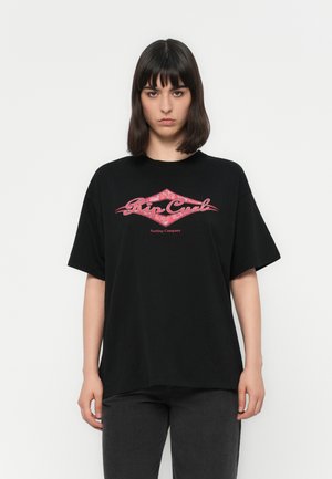 Woman with shoulder-length dark hair wearing a black Rip Curl Surfing Company T-shirt and dark pants, standing against a white background.