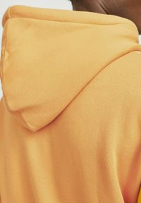 Oranje hoodie met een zachte textuur, voorzien van een ronde trekkoordkap en een strakke naaddesign. De stof lijkt lichtgewicht en comfortabel.
