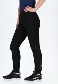 Black athletic pants with a fitted design, zippered pockets, and contrasting gold accents. Made of stretchy, textured fabric.