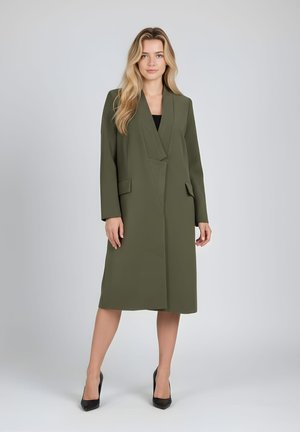 Mujer rubia de pie, con abrigo verde oliva hasta la rodilla sobre camiseta negra y tacones altos negros, contra un fondo gris claro liso.
