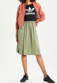 Jasnoróżowa kurtka bomberka z prążkowanymi mankietami, czarna koszulka z białym logo Adidas, zielona spódnica do kolan i czarne trampki.