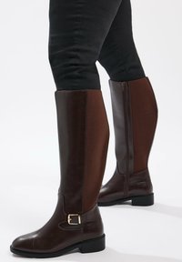 Bruna knähöga stövlar med en texturerad brun panel, framknäppningsdetalj och sidodragkedja. Slät lädermaterial med låg klack.