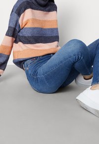 Pletený pruhovaný svetr v růžové, broskvové, námořnické a šedé barvě; spárován s modrými skinny džíny a bílými teniskami, sedící na šedém pozadí.