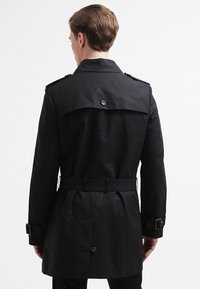Czarny płaszcz trench z paskiem w talii, epoletami na ramionach oraz guzikowym klapą z tyłu. Wykonany z gładkiego, wodoodpornego materiału.