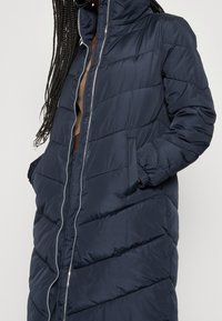 Námořnický puffer kabát s prošívaným designem, vysokým límcem a zipovým zavíráním. Má boční kapsy a elastické manžety pro těsné padnutí.