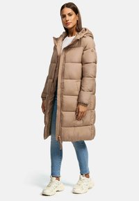 Long, puffer coat in beige with a hood, quilted design, front zipper, and elastic cuffs, paired with light blue jeans and beige sneakers.