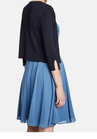 Marinblå kort cardigan med trekvartsärmar, stickad textur, som bärs över en blå klänning med utsvängd kjol och svaga plisseringar.