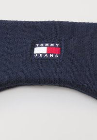 Tumedadest kudumine kõrvakattega, millel on ristkülikukujuline plaastriga, kus on valge, punase ja sinise värviga "TOMMY JEANS" logo. Pehme tekstuur, elastne istuvus.