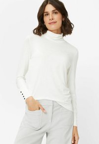 Langærmet hvid rullekrave med en glat tekstur og subtil glans, prydet med knapdetaljer ved manchetterne, som er parret med grå bukser.