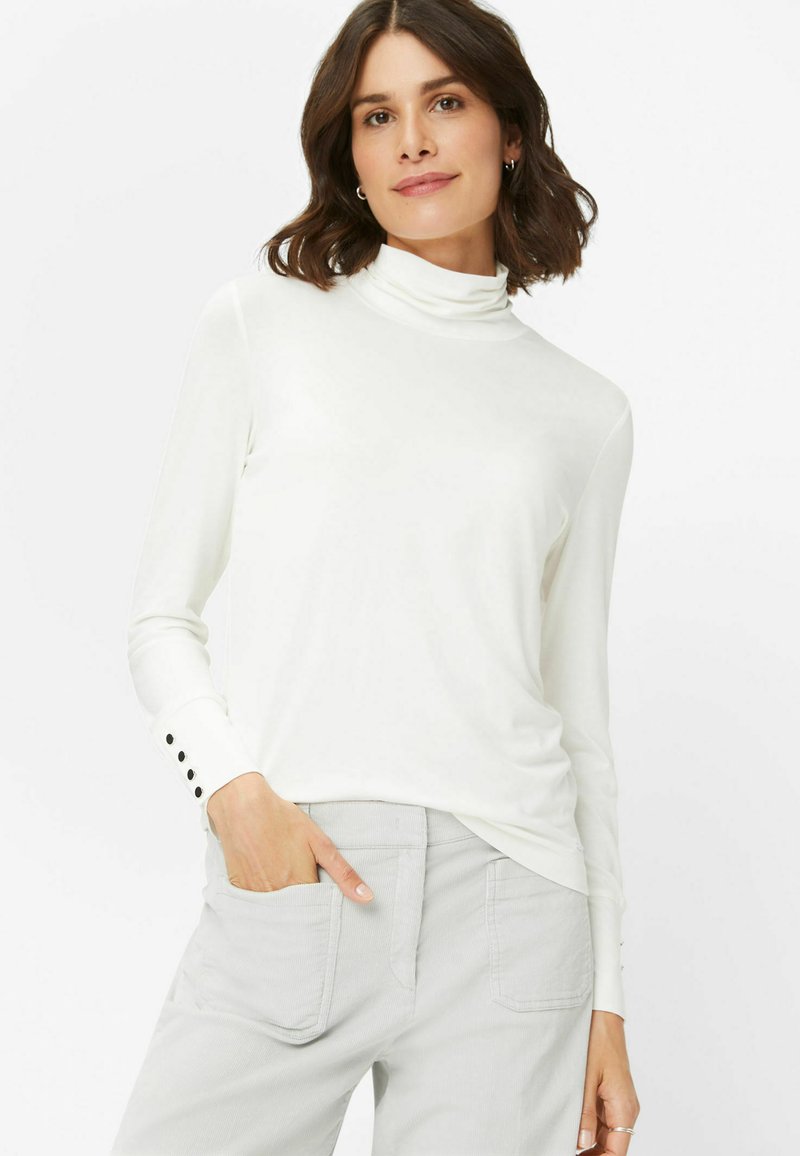 Langærmet hvid rullekrave med en glat tekstur og subtil glans, prydet med knapdetaljer ved manchetterne, som er parret med grå bukser.