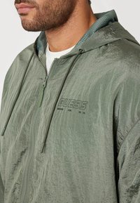 Zöld, könnyű dzseki strukturált felülettel, cipzáras elülső résszel és állítható kapucnival. Hímzett "GUESS" logóval és kontrasztos hálós béléssel rendelkezik.