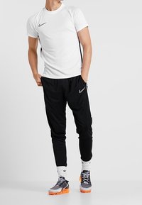 Camiseta blanca de manga corta de Nike, pantalones deportivos negros con franjas laterales y zapatillas Nike negras y naranjas estampadas, con puños acanalados.
