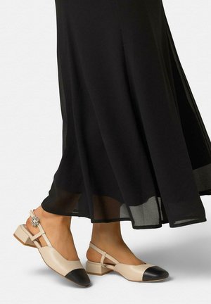 Pies con zapatos slingback beige con punteras negras y hebilla decorativa, combinados con un vestido negro transparente y fluido hasta el tobillo.