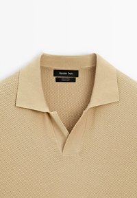 Polo shirt in cotone beige con un motivo testurizzato, colletto a V e orlo a coste. L'etichetta indica "Massimo Dutti, 100% Cotone, Prodotto in Cina."