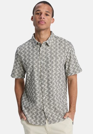 Mann trägt ein kurzärmeliges, geknöpftes Hemd mit schwarzem und weißem, vertikalem abstraktem Muster und hellfarbene Hose, Hände in den Taschen.