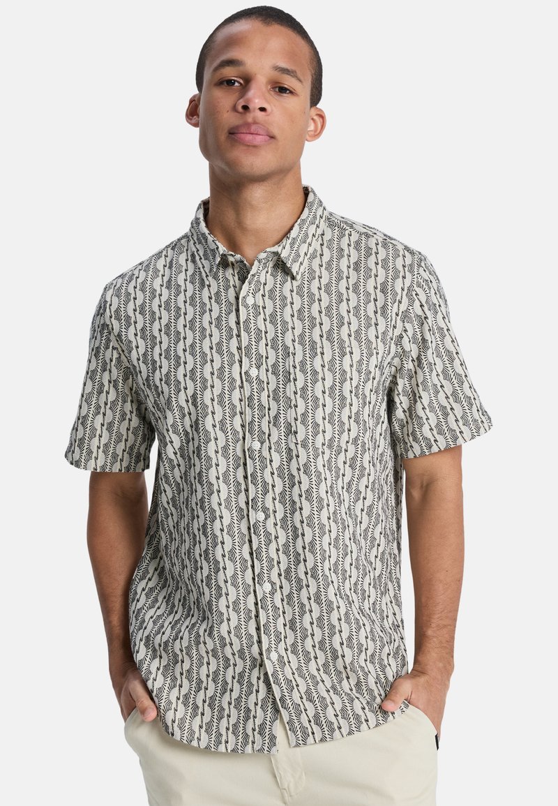 Άνδρας με κοντομάνικο πουκάμισο με μαύρο και λευκό κάθετο αφηρημένο μοτίβο και ανοιχτόχρωμο παντελόνι, με τα χέρια στις τσέπες.