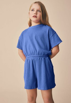 Mädchen mit langen blonden Haaren, das ein blaues kurzärmeliges Oberteil und passende Shorts mit elastischem Bund trägt und vor einem beigen Hintergrund steht.