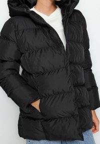 Czarna kurtka puchowa z kapturem, z pikowanym wzorem, gładką teksturą i dwoma bocznymi kieszeniami. Stylizowana z białym swetrem pod spodem.