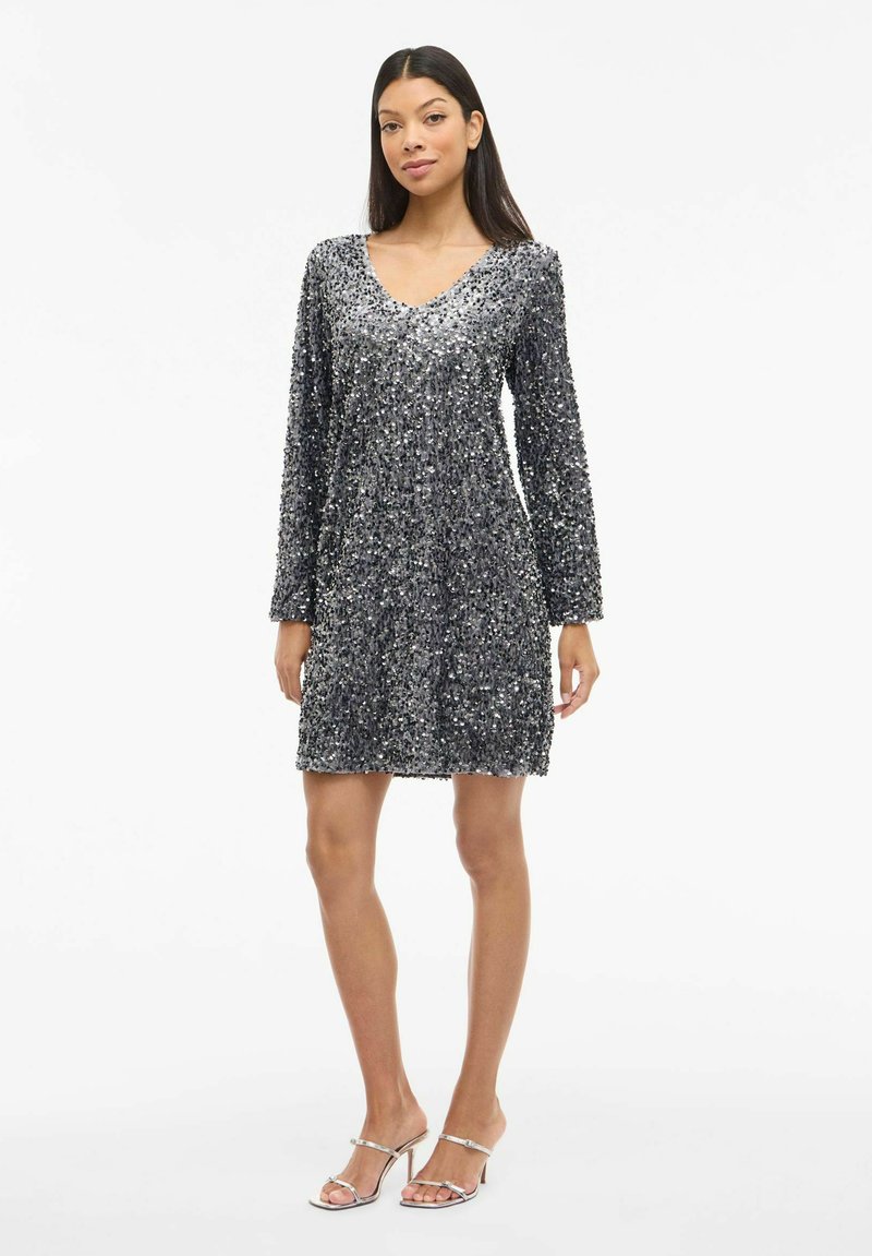 Sudraba pailletēta kleita ar V-veida izgriezumu, gariem piedurknēm un brīvu piegriezumu, kas atspoguļo gaismu. Kombinēta ar sudraba siksnām rotātiem augstpapēžu apaviem.